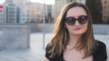 Portrait of young businesswoman in sunglasses walking in city street. Attractive business woman looking at camera. Face of confident girl commuting to work. Blurred background. Close up Slow motion.