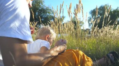 Çim, sarı saçlı Sevimli oğluyla bir güneşli yaz gününde oturan genç baba profil. Mutlu bir aile birlikte doğa zaman harcama. Arka plan, güzel doğası. Ağır çekim yakın çekim.