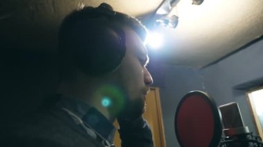 Profile of male singer in headphones singing song into the microphone at sound studio. Young man recording new melody or song. Working of creative musician. Show business concept. Slow motion.