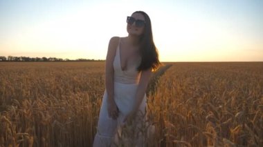 Beautiful brunette standing among wheat field showing joyful emotions. Smiling girl having fun outdoor playing with her hair. Happy woman enjoying summer with sunset at background. Slow motion.