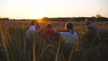Mommy and daddy with children sitting on grass at field and hugging each other at sunset. Young parents with two kids spending time together at meadow. Concept of family care and love. Dolly shot.