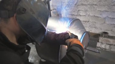 Atölyede koruyucu maske kaynakçısı. İki çelik detay arasında ortak çalışma yapan bir işçi. Adam metal parçalarını birbirine kaynaştırıyor. Metalle el emeği. Dolly vurdu..