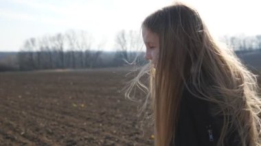 Beautiful small girl with long blonde hair walks on the dry ground through ploughed field at early spring. Happy little child in jacket goes on the dirty soil over plowed plantation. Close up.