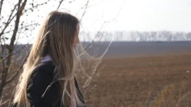 Little smiling girl with backpack goes along a driveway near ploughed meadow. Small kid moves on way to school at spring day. Happy female child with long blonde hair walks on road near plowed field.