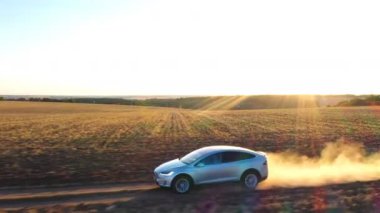 Arazi yolundan geçen elektrikli arabanın hava görüntüsü ardında toz izi bırakıyor. Kırsal yol boyunca giden elektrik yükündeki ekoloji dostu otomobil. Arkaplanda manzaralı günbatımı manzarası.