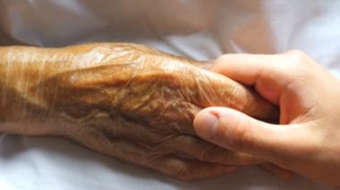 Worried man holding and gently stroking hand of his sick grandmother showing care or love. Son comforting wrinkled arm of elderly mom lying at bed. Guy giving support to his old parent. Top detail