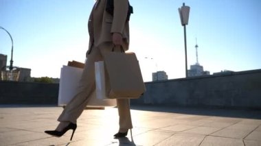 Elegant young lady holds shopping bags going at city square after purchases. Attractive woman carries paper packets walking along sunny urban street. Sales and discounts concept. Slow motion.