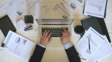 Male hands of manager typing text on laptop keyboard at white desk and drinking coffee. Businessman working on notebook at wooden table in office. Diagrams with statistics at background. Top view.