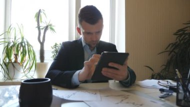 Young entrepreneur analyzing statistical data information on a tablet pc in office. Serious businessman looking at diagrams with statistics on paper while working on digital device. Dolly shot.