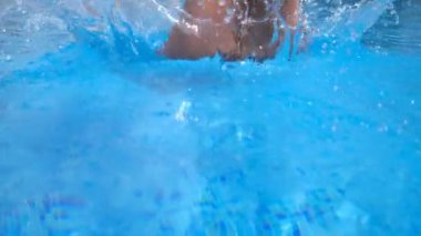 Close up of young woman diving in pool and splashing crystal clear water. Girl in swimsuit swimming in basin and enjoying holiday. Lady relaxing during summer vacation. Low angle view Slow motion.