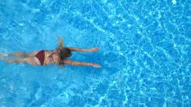 Young tanned woman swimming in pool with transparent clear water at sunny day. Unrecognizable sexy girl enjoying her summer vacation on resort floating in basin. Concept of travel. Slow mo Top view.
