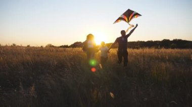 Mother and father with small daughter holds hands of each other running among summer meadow at sunset time. Young parents with little cute girl jogging with rainbow kite through grass field. Close up.
