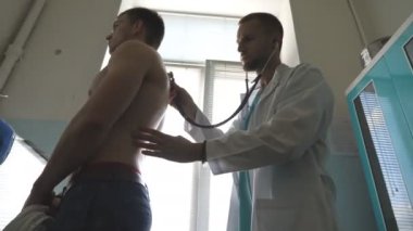 Doktor steteskoplu genç bir erkek hastayı muayene ediyor. Doktor, hastanedeki ofisteki adamın arkasını kontrol ediyor. Atletik adamın kalp atışlarını dinleyen bir tıp çalışanı. Kapat..