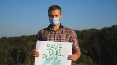 Gezegeni kurtarmak için çevreci hareketin posterini tutan genç adamın portresi. Koruyucu maskeli bir erkek ekolojik aktivist kameraya bakıyor ve doğanın korunmasını savunuyor. Kapat..