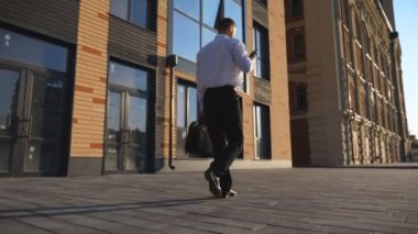 Başarılı bir iş adamı resmi gözlük takıyor cep telefonuyla kaldırımda işe gidiyor. Evrak çantalı genç yönetici şehir caddesindeki ofis binasının yakınında akıllı telefon kullanıyor. Dolly çekim ağır çekim.