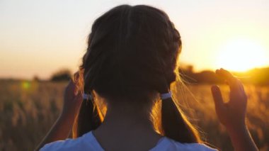 Sevimli küçük kız çayırda duruyor sarı saçlarını düzleştiriyor. Güzel bayan çocuk kameraya dönüp gülümsüyor. Yaz tarlasında küçük mutlu bir çocuğun portresi. Bulanık gün batımı arkaplanı.