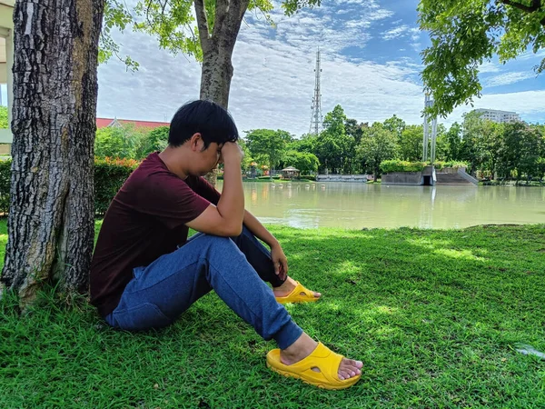 Young Asian man with black hair sitting under a tree. Stress and anxiety. What are you thinking in the summer park city