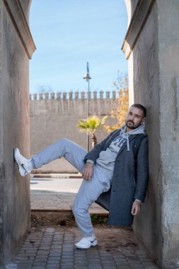 Faslı bir adam duvara yaslanmış şık gri bir ceket ve beyaz Nike ayakkabılar giyiyor.