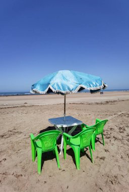 Fas sahilinde yeşil sandalyeli ve mavi şemsiyeli bir masa.