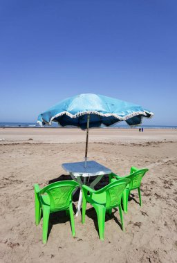 Fas sahilinde yeşil sandalyeli ve mavi şemsiyeli bir masa.