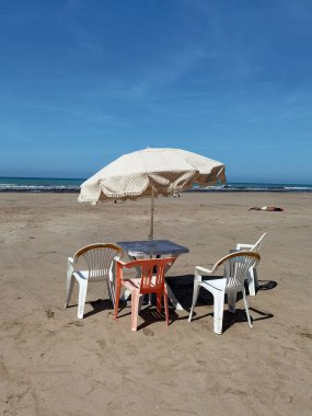 Beyaz sandalyeli bir masa ve Fas sahilinde beyaz bir şemsiye.