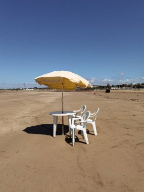 Beyaz sandalyeli bir masa ve Fas sahilinde sarı bir şemsiye.