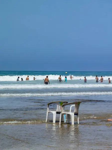 Sahilde iki beyaz sandalye ve okyanusta yüzen yerli halk.