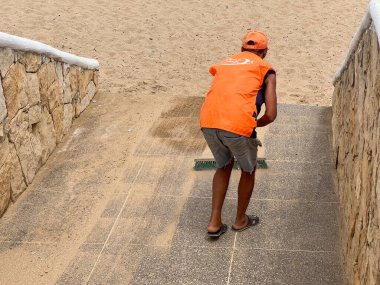 Temizlikçi, kumsala giden yolu manuel süpürgeyle süpürüyor.