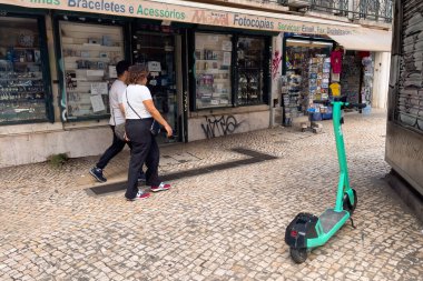 Lizbon caddesinde park etmiş bir E-scooter 'ın yanından geçen insanlar.