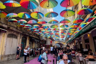 Lizbon 'daki binaların arasında asılı renkli gökkuşağı şemsiyeleriyle pembe sokakta takılan turistler.