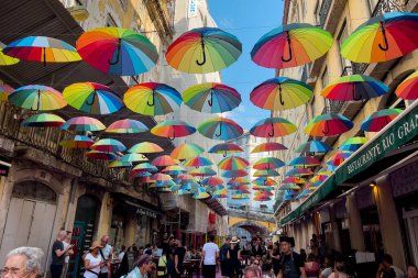 Lizbon 'daki binaların arasında asılı renkli gökkuşağı şemsiyeleriyle pembe sokakta takılan turistler.