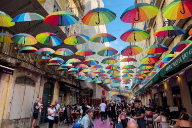 Lizbon 'daki binaların arasında asılı renkli gökkuşağı şemsiyeleriyle pembe sokakta takılan turistler.