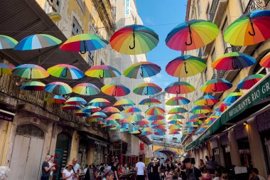 Lizbon 'daki binaların arasında asılı renkli gökkuşağı şemsiyeleriyle pembe sokakta takılan turistler.