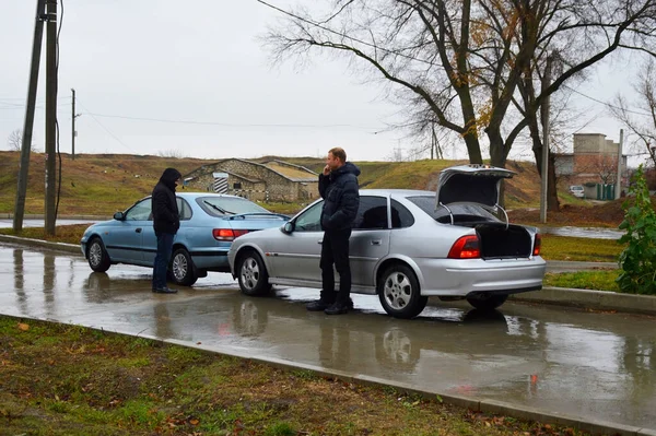 two cars after an accident - a collision on a wet road, drivers are standing nearby. one looks confused at the impact site, the other calls the insurance agency and the police