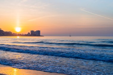 Sahilde gün doğumu, dağın arkasında doğan güneş, arka planda küçük bir tekne, denizin dalgaları, tepede uçak.