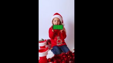 Noel zamanı. Küçük bir kız elinde yeşil ekranlı, Noel Baba şapkalı, kırmızı kazaklı, süslü püslü ve taşaklı bir telefon tutuyor. Kavram reklamcılığı, teknoloji, duyuru, tebrikler, davetiyeler, modeller..