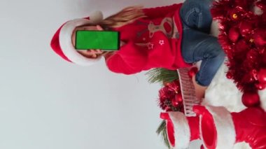 Noel zamanı. Küçük bir kız elinde yeşil ekranlı, Noel Baba şapkalı, kırmızı kazaklı, süslü püslü ve taşaklı bir telefon tutuyor. Kavram reklamcılığı, teknoloji, duyuru, tebrikler, davetiyeler, modeller. 
