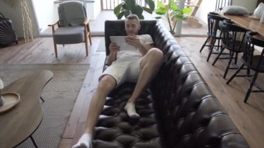 Elinde tablet olan bir adam evinde kanepede dinleniyor.