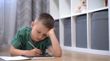 Bir çocuk telefonundan video izliyor ve odasında yerde yatarken not defterine not alıyor.