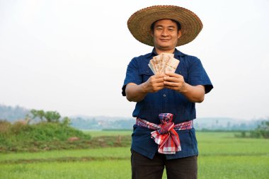 Asyalı çiftçinin portresi Tayland paralarını çeltik tarlasında tutuyor. Mutlu çiftçi kar, gelir ve tarım desteği alıyor. Ekinlerle gurur duyuyorum..      