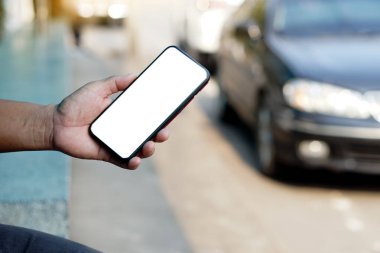 Elleri beyaz beyaz ekranlı akıllı telefonu tutuyor. Arabanın arka planı bulanık. Konsept: Araba servisi ve ulaşım teknolojisi için akıllı telefonda mobil internet uygulamaları kullanmak.     