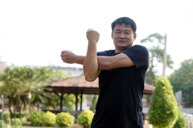 Asyalı bir adamın portresi parkta egzersiz yapıyor. Omuz esnemesi. Sağlıklı yaşam tarzı kavramı. Tam vücut esneme hareketi. Yağları ve kalorileri yakın. Egzersiz fiziksel ve zihinsel sağlık için iyidir..