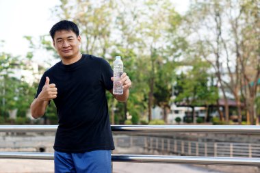 Asyalı adam parkta duruyor, elinde bir şişe içme suyu tutuyor ve parmağını kaldırıyor. Kavram: sağlık için su iç. Sağlıklı yaşam tarzı.  