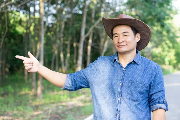 Yakışıklı Asyalı çiftçi şapka takıyor ve mavi gömlek orman bahçesinde, parmakla işaret ediyor, kendine güven duyuyor, orman bahçesinde duruyor. Kavram: ormancı. Orman koruma. Bahçe olarak büyüyen ekonomi ağaçları