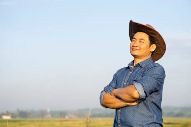Yakışıklı Asyalı çiftçi şapka takıyor, kollarını göğsüne kavuşturup gülümsüyor. Kendinden emin hissediyor. Konsept: mutlu çiftçi. Tarım işgali. Metin veya reklam eklemek için boşluk kopyala.