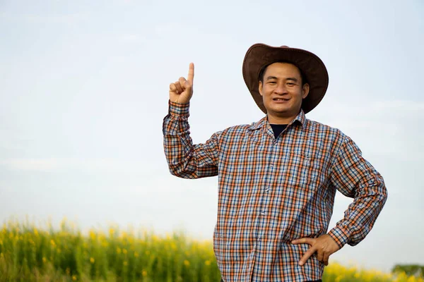 Portrait of handsome Asian man farmer is at garden, wears hat, plaid shirt, raise finger point up to sky, hand touch on hip. Concept : Happy Thai farmer. Copy space for adding text or advertisement.