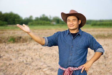 Yakışıklı Asyalı çiftçinin portresi tarımda, şapka takıyor, mavi gömlek giyiyor, beline Tayland peştemali takıyor, mevcut ürünü gösteriyor, kameraya bakıyor. Konsept: mutlu çiftçi.