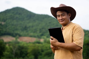 Yakışıklı Asyalı botanikçi doğa parkında elinde akıllı tablet tutuyor. Kameraya bak, bulanık dağ arkaplanı. Kavram: Kablosuz teknolojiyi araştırmak, doğal kaynakları araştırmak için kullanmak. 