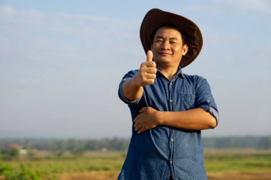 Asyalı çiftçinin portresi şapka takıyor, başparmak yukarı. Gülümsüyor. Kendinden emin hissediyor. Konsept: mutlu çiftçi. Tarım işgali. Metin veya reklam eklemek için boşluk kopyala.