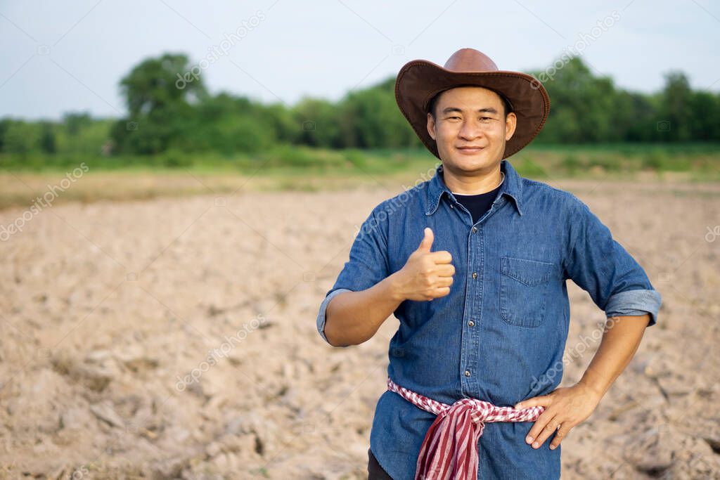 Handsome Asian man farmer is at plantation. He wears hat, blue shirt, Thai loincloth on waist, put hand on hips, thumbs up, feels confident, looks at camera. Concept : Agriculture occupation. 